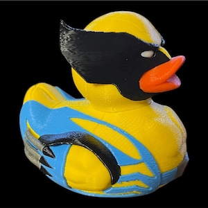 May include: A yellow rubber duck designed to resemble a superhero character, featuring a black mask over the eyes, an orange beak, and a blue suit pattern on the body. The figure includes black wing-like accents and silver claw-like protrusions.