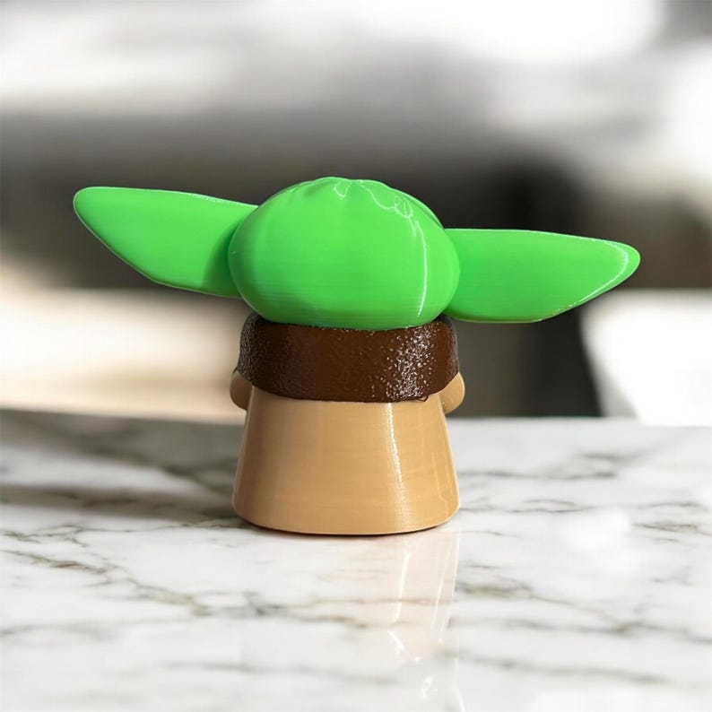 May include: A small, green and brown figurine of a character from the Star Wars franchise. The character is wearing a brown robe and has large, green ears.