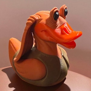 Op de afbeelding: Oranje en groene badeend die lijkt op het Star Wars-personage Jar Jar Binks. De eend heeft grote ogen, lange oren en een brede, oranje snavel. Hij draagt een groen vest.