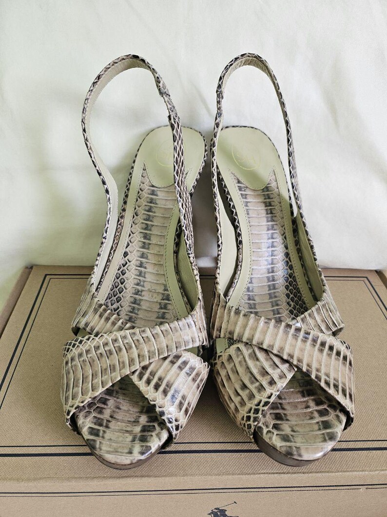 May include: A pair of open-toe slingback sandals with a snakeskin pattern in beige, brown, and black. The sandals feature a crisscross design and a thin ankle strap. The heels are approximately 7.6 cm high.