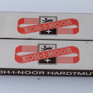 Puede incluir: Dos cajas blancas apiladas una encima de la otra. Las cajas están etiquetadas con el texto "KOH-I-NOOR" en letras rojas y negras. La caja inferior también tiene el texto "HARDTMUTH" en letras negras.