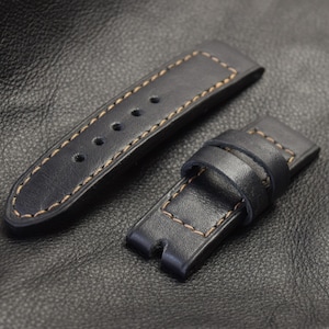 Puede incluir: Dos correas de reloj de cuero negro con costuras de color naranja contrastantes. Las correas están dispuestas planas sobre una superficie de cuero negro.