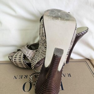 May include: Snakeskin-patterned high-heeled sandals in brown, beige, and black. The dark brown heels have a curved design. The soles are off-white. The sandals are on a cardboard box. The shoe size is 40.