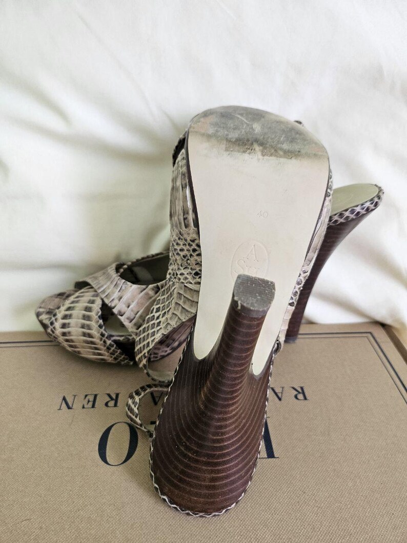 May include: Snakeskin-patterned high-heeled sandals in brown, beige, and black. The dark brown heels have a curved design. The soles are off-white. The sandals are on a cardboard box. The shoe size is 40.