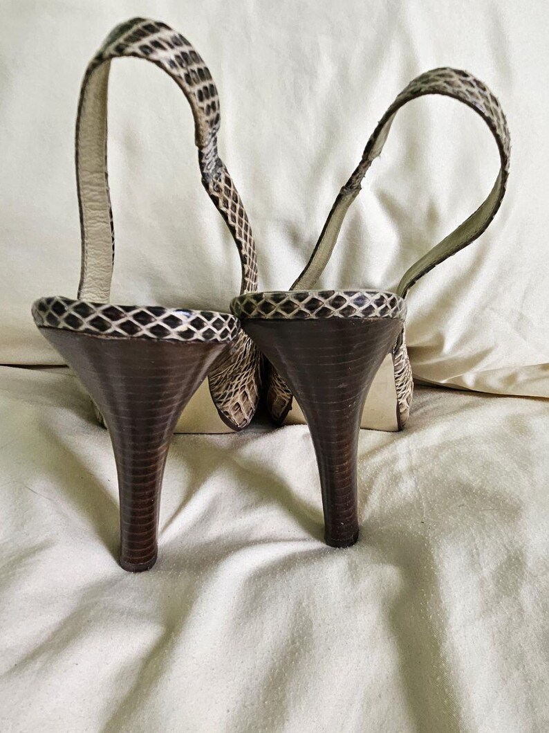 May include: A pair of high-heeled sandals with a snakeskin pattern. The heels are dark brown and tapered. The straps are thin and loop around the ankle. The sandals are set against a neutral background.