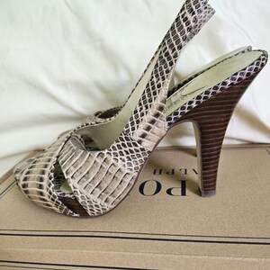 May include: Snakeskin-patterned slingback sandals with high heels in beige and brown. The dark brown heels are set on a cardboard box. The sandals have a strap around the ankle.