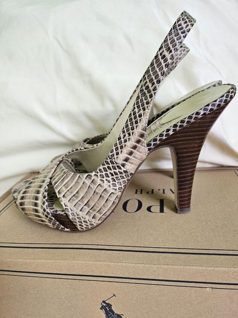 May include: Snakeskin-patterned slingback sandals with high heels in beige and brown. The dark brown heels are set on a cardboard box. The sandals have a strap around the ankle.