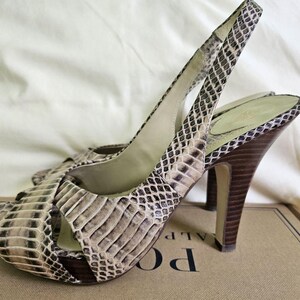 May include: A pair of open-toe slingback high heels with a snakeskin pattern in shades of beige, brown, and black. The heels are dark brown and the shoes are resting on a cardboard box.