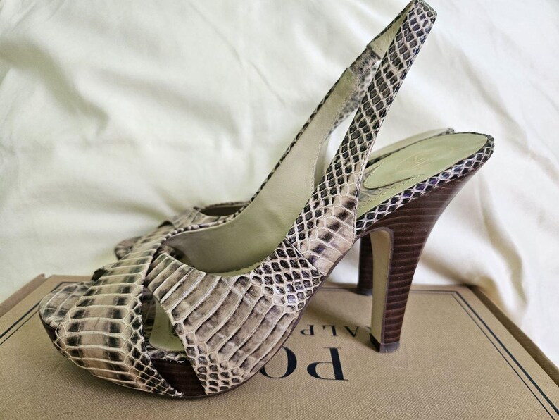 May include: A pair of open-toe slingback high heels with a snakeskin pattern in shades of beige, brown, and black. The heels are dark brown and the shoes are resting on a cardboard box.