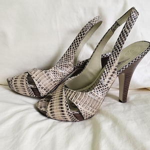 Può includere: Un paio di sandali slingback con tacco alto e motivo a pelle di serpente nei toni del beige, marrone e nero. I tacchi sono in legno e i sandali hanno la punta aperta.