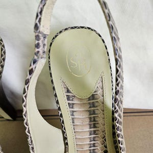 May include: Close-up of a snakeskin-patterned sandal. The sandal features a slingback design with a high heel and a light-coloured insole. The insole has a gold logo. The shoe's pattern includes shades of brown, beige, and black.