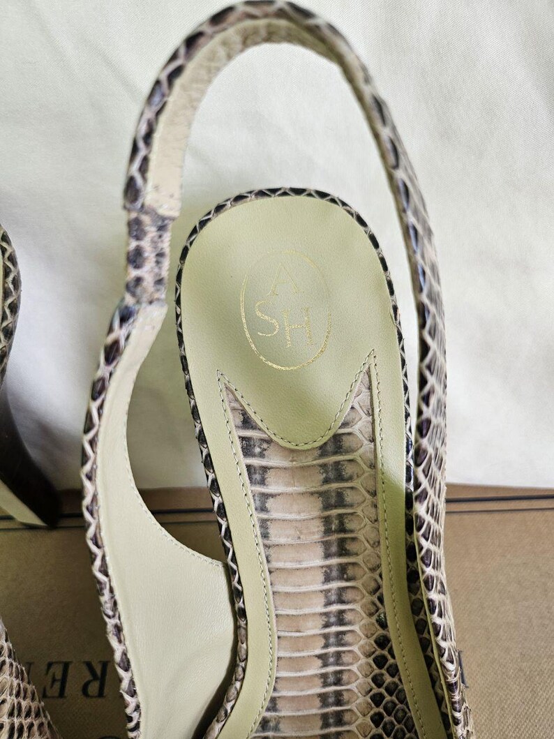 May include: Close-up of a snakeskin-patterned sandal. The sandal features a slingback design with a high heel and a light-coloured insole. The insole has a gold logo. The shoe's pattern includes shades of brown, beige, and black.