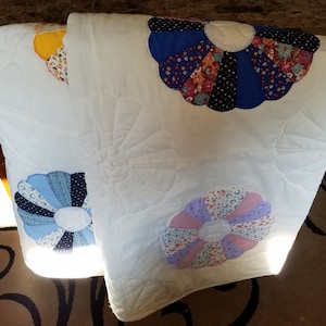 May include: A white quilt featuring three large, colourful fabric flower appliques. The flowers are in shades of blue, purple, and yellow. The quilt is folded in half and lying on a table.