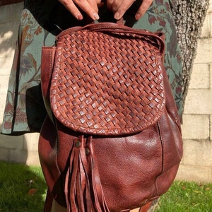 May include: A brown leather backpack with a woven flap and a long tassel. The backpack has a top handle and adjustable straps.