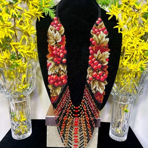 May include: A beaded necklace with a black cord and a cascade of red, gold, and black beads. The upper portion features a leaf and berry design in red, gold, and brown. The necklace is displayed on a black mannequin.