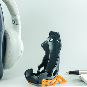 May include: A black and orange 3D printed racing seat model. The seat is small and appears to be a miniature version of a real racing seat. The seat is sitting on a white surface.