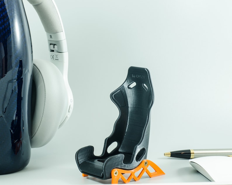 May include: A black and orange 3D printed racing seat model. The seat is small and appears to be a miniature version of a real racing seat. The seat is sitting on a white surface.