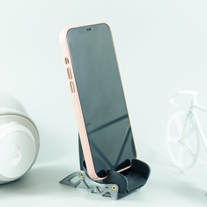 May include: A black phone stand with a rose gold phone in it. The phone is on a white surface.