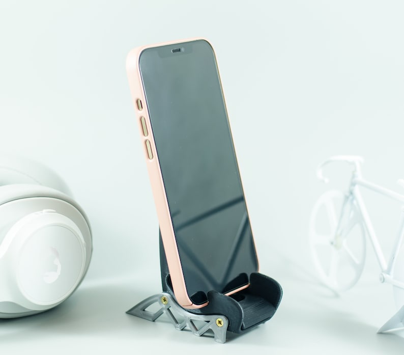 May include: A black phone stand with a rose gold phone in it. The phone is on a white surface.
