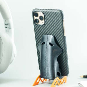 May include: A black phone case with a carbon fiber pattern is in a black phone stand. The phone stand is on an orange stand with a geometric design.
