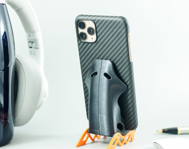 May include: A black phone case with a carbon fiber pattern is in a black phone stand. The phone stand is on an orange stand with a geometric design.