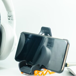 May include: A black smartphone is held in a black and orange phone stand. The phone is in portrait mode and the screen is black. A white pair of headphones are in the background.