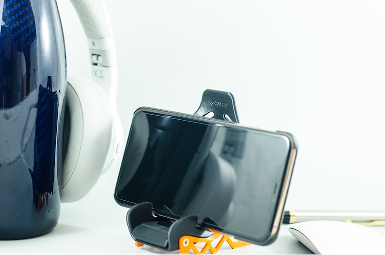 May include: A black smartphone is held in a black and orange phone stand. The phone is in portrait mode and the screen is black. A white pair of headphones are in the background.
