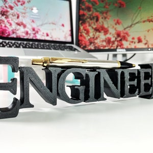 May include: Black 3D printed word "Engineer" on a white surface with a gold pen in front of it.