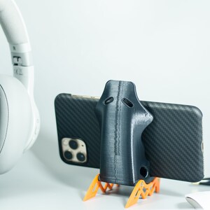 May include: A black 3D printed phone stand with orange legs, holding a black smartphone with a carbon fiber case. The phone stand is designed to hold the phone upright.