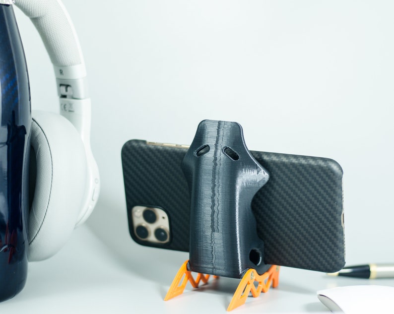 May include: A black 3D printed phone stand with orange legs, holding a black smartphone with a carbon fiber case. The phone stand is designed to hold the phone upright.