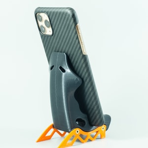 May include: A black phone stand with a carbon fiber pattern on the back. The stand has a black phone holder and orange legs.