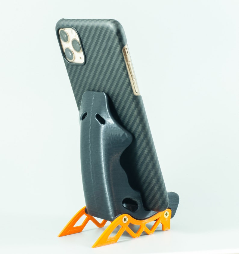 May include: A black phone stand with a carbon fiber pattern on the back. The stand has a black phone holder and orange legs.