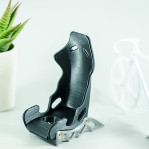 May include: A black 3D printed racing seat model with a silver metal base. The seat is designed with a high back and side bolsters for support. The model is sitting on a white surface.