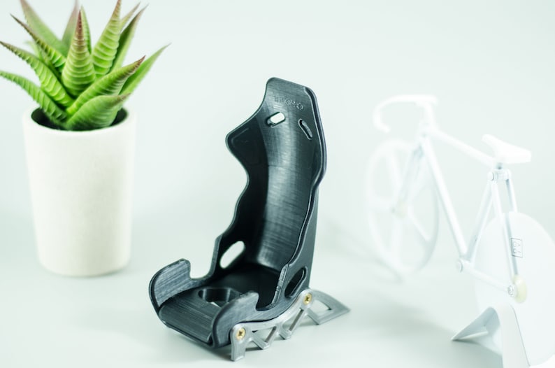 May include: A black 3D printed racing seat model with a silver metal base. The seat is designed with a high back and side bolsters for support. The model is sitting on a white surface.