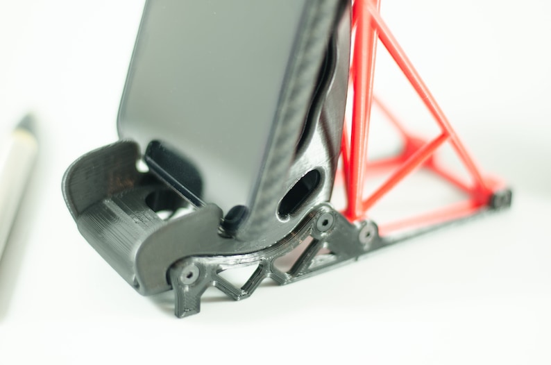 Może przedstawiać: Czarny stojak na telefon z czerwoną, tr&oacute;jkątną podstawą. Stojak jest wykonany z plastiku drukowanego w 3D i ma fakturowaną powierzchnię.