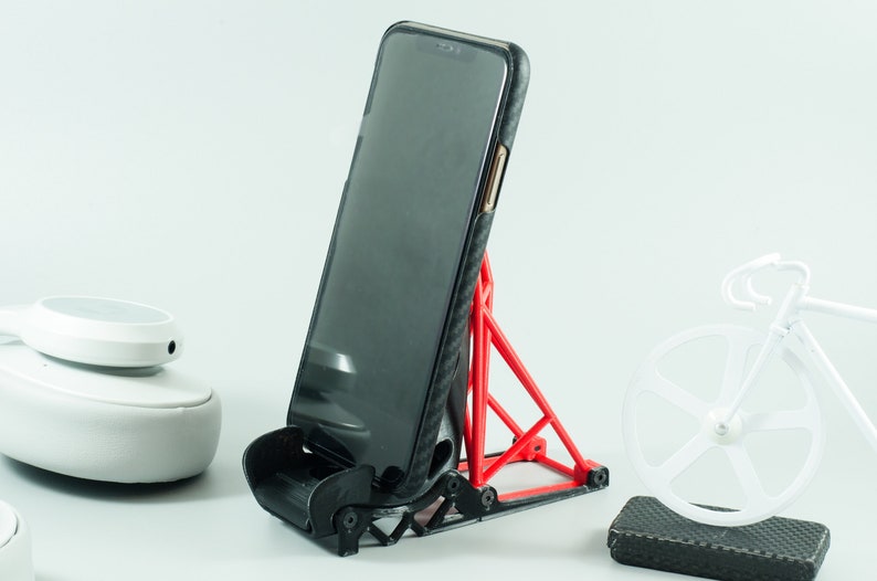 Może przedstawiać: Czerwony stojak na telefon, wydrukowany w 3D, z czarnym smartfonem w czarnym etui. Stojak został zaprojektowany z tr&oacute;jkątną podstawą i zakrzywionym wierzchołkiem, aby utrzymać telefon w pozycji pionowej. Stojak znajduje się na białym tle.