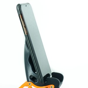 May include: A black phone case with a carbon fiber pattern is in a black and orange phone stand. The phone is in portrait mode.