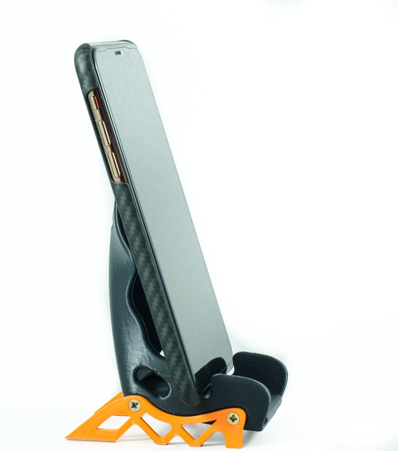 May include: A black phone case with a carbon fiber pattern is in a black and orange phone stand. The phone is in portrait mode.