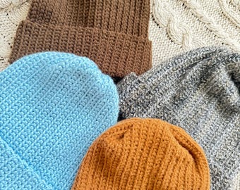 May include: Four knitted hats in different colors: brown, light blue, brown and gray. The hats are arranged in a pile on a white knitted surface.