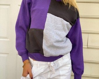 May include: A long-sleeve crewneck sweatshirt featuring a color-blocked design in purple, black, and light gray. The garment is paired with white cargo-style pants that include a drawstring waist and a small metal ring detail on the front pocket.