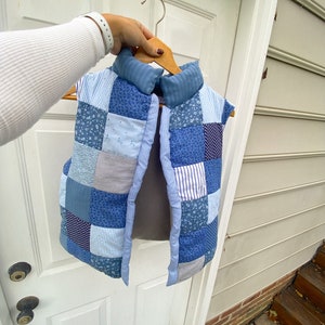 May include: A patchwork vest in shades of blue, held on a wooden hanger. The vest features a variety of blue fabrics, including floral, striped, and solid patterns. The collar is a solid blue fabric. The vest is open in the front.