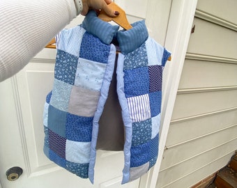 May include: A patchwork vest in shades of blue, held on a wooden hanger. The vest features a variety of blue fabrics, including floral, striped, and solid patterns. The collar is a solid blue fabric. The vest is open in the front.