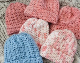 May include: A collection of handmade crochet beanies in various colors. The hats are primarily pink, with one blue and two white and pink speckled designs. The beanies are arranged on a textured, light gray surface.