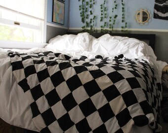 May include: A bed covered with a black and white checkered quilt. The quilt is draped over white sheets and pillows. The room has light blue walls, a window, and decorative plants.