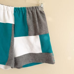 May include: Shorts featuring a color-blocked patchwork design in solid white, teal, and heather gray fabric panels. The garment has an elasticized waistband and is displayed on a clear plastic hanger, showcasing a multi-paneled construction.