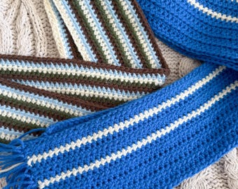 May include: Handmade crochet scarves in shades of blue, white, brown, and green. One scarf is solid blue with white stripes, and another features a striped pattern. The scarves have fringed ends and are displayed on a textured surface.
