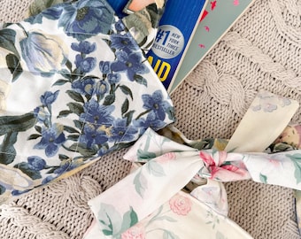 May include: Two floral fabric tote bags with tied handles. One bag features blue flowers on a white background, and the other has pink flowers. Books are visible inside one of the bags, including a New York Times bestseller.