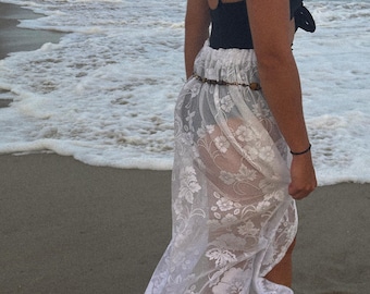 May include: A white lace maxi skirt with a floral pattern. The skirt is sheer and has a slit up the side. The skirt is being worn by a person on a beach.