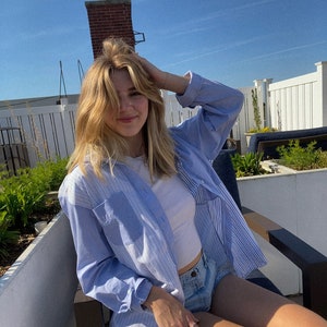 May include: A woman wearing a light blue striped button-down shirt over a white tank top and denim shorts. She is sitting on a chair with her hand in her hair.