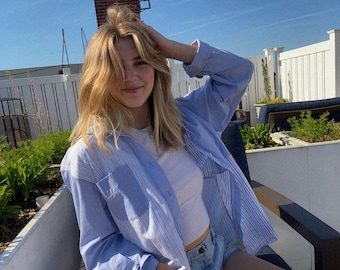 May include: A woman wearing a light blue striped button-down shirt over a white tank top and denim shorts. She is sitting on a chair with her hand in her hair.